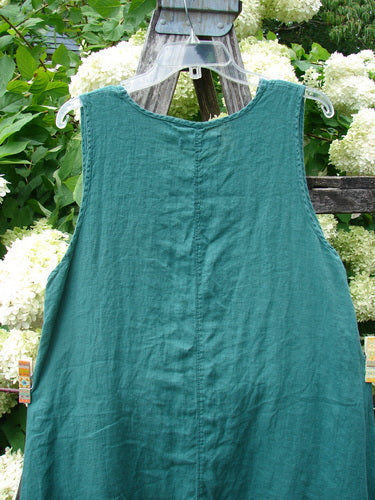 Barclay Linen Round Bottom Pinafore in Cloud Burst Jade, Size 0, a sleeveless linen dress with a gently rounded hem, center seam, and soft wrinkled texture, hanging on a clear hanger against white flowers and green foliage.