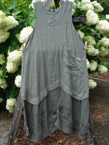 Barclay Linen Explorer Jumper in Buttercup Green River, size 0, a sleeveless linen dress with a soft gray-green, crinkled texture, layered asymmetrical hem, and a gathered front tie. It hangs outdoors against white hydrangeas and dark wood, with muted natural light.