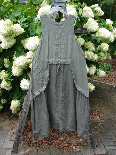 Barclay Linen Explorer Jumper in Buttercup Green River, size 0, is a sleeveless linen garment with a soft, crinkled texture and layered drape. It hangs from a wooden stand outdoors, framed by white hydrangeas and muted garden greenery.
