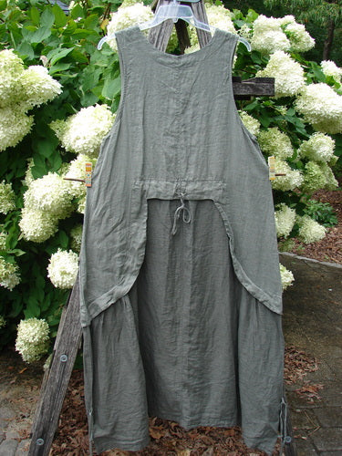 Barclay Linen Explorer Jumper in buttercup green, size 0, hangs from a weathered wooden ladder amid dense white hydrangeas. The sleeveless linen piece has a softly crinkled texture, a loose draped silhouette, and a gathered front tie that adds structure to the muted green-gray fabric.