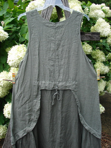 Barclay Linen Explorer Jumper in Buttercup Green River, size 0, with a sleeveless A-line silhouette, soft linen texture, and a gathered tie detail at the lower front. The muted green garment hangs against leafy white hydrangeas, creating a natural, layered backdrop.
