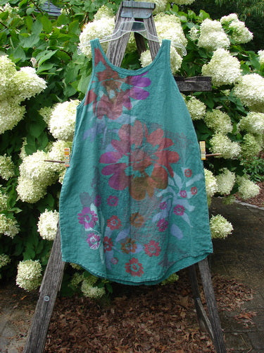 Barclay Linen Round Bottom Pinafore Giant Floral Jade Size 0, a teal linen pinafore with a rounded hem and thin shoulder straps, printed with oversized floral motifs in purple, pink, and rust. It hangs from a weathered wooden ladder amid white hydrangeas, with soft outdoor light and earthy greens framing the scene.