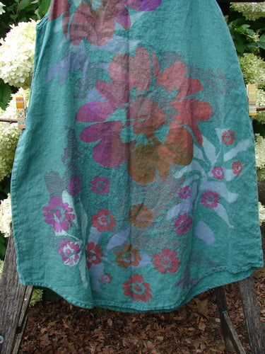 Barclay Linen Round Bottom Pinafore Giant Floral Jade Size 0 in sheer jade linen, with a rounded hem and oversized floral print in purple, pink, and teal. The fabric hangs in a soft, layered drape against an outdoor garden backdrop.