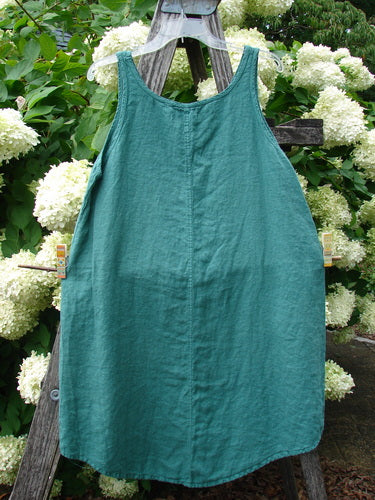 Barclay Linen Round Bottom Pinafore Giant Floral Jade Size 0 in teal-green linen, with a sleeveless, round-bottom silhouette and softly crinkled texture. It hangs centered on a wooden ladder outdoors, framed by dense white hydrangea blooms and leafy greenery in natural light.