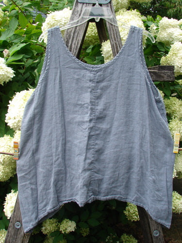 Barclay Linen Altoll Top Pansy Patch Blue Dust size 0, a loose sleeveless linen top in faded blue-gray with a wide scoop neckline, raw-looking edges, and a softly wrinkled drape hanging from a wooden frame amid pale flowers.