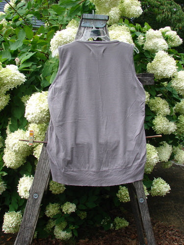 Barclay Flutter Bottom Tank Top in Moon Bird stone plum, size 2, shown as a sleeveless gray tank with a softly flared hem and smooth, lightweight knit texture, hanging centered on a weathered wooden ladder among dense white hydrangeas and green foliage.