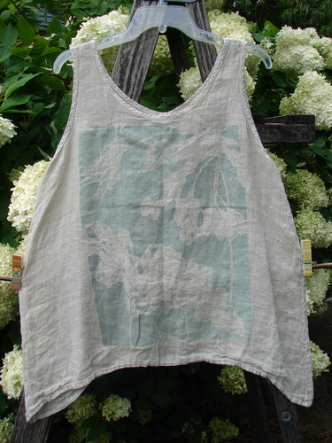 Barclay Linen Altoll Top Blossom Reach Creme, size 0, is a sleeveless linen top in soft cream with a faded pale blue floral graphic across the front. The crinkled, airy fabric hangs from a clear hanger, its relaxed scoop neckline and uneven hem framed by lush white hydrangeas and dark wood outdoors.
