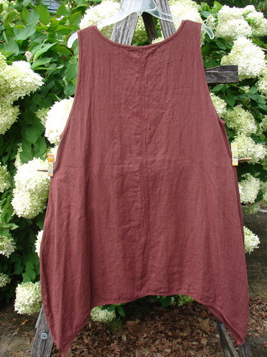 Barclay Summercloth Round Pocket Pinafore Star Flower in rusty bucket size 0, a sleeveless rust-colored linen pinafore with a rounded neckline and soft drape. It hangs centered against white hydrangeas and weathered wood, with a muted, earthy finish.
