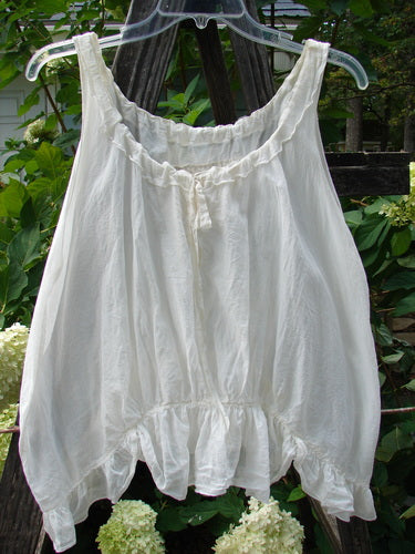 Magnolia Pearl Silken Cotton Voile Ribbon Neck Top White OSFA hanging on a clothesline, showcasing its delicate fabric, adjustable ribbon neckline, tiny pearl buttons, and varying hemline.