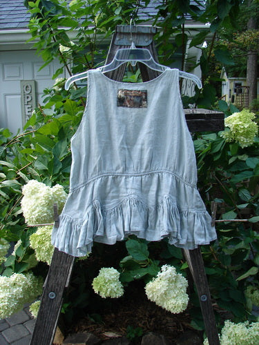 Magnolia Pearl Linen Roman Ruffle Top Light Sage OSFA displayed on a wooden ladder adorned with white flowers, showcasing its rounded neckline, ruffled hemline, and signature patch.