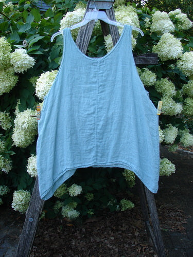Barclay Linen Altoll Top Falling Floral Sage size 2 in pale sage linen, a sleeveless, loose-fitting top with a wide scoop neckline and softly draped hem. It hangs from a wooden stand, its airy texture set against dense white hydrangeas and green foliage.