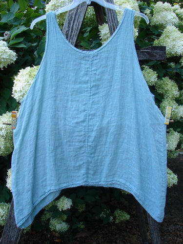 Barclay Linen Altoll Top Falling Floral Sage Size 2 in sage linen, a loose sleeveless top with a wide scoop neckline, softly textured drape, and uneven hem, photographed hanging against leafy white hydrangeas and dark wood.