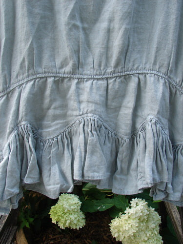 Magnolia Pearl Linen Roman Ruffle Top Light Sage OSFA, featuring a rounded neckline, scalloped ruffled hemline, and signature patch, captured up close with delicate white flowers in the background.