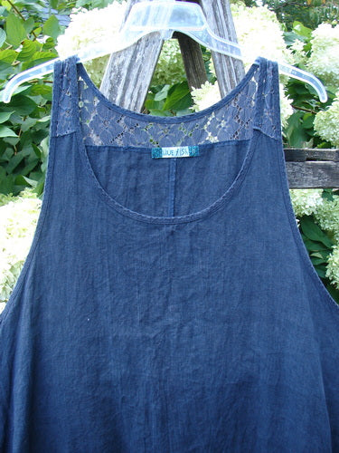 Barclay Summercloth Lace Two Pocket Pinafore in unpainted black, size 2, with a sleeveless A-line shape, lace inset at the neckline, and a soft denim-like texture. It hangs on a clear plastic hanger outdoors, with green foliage behind it.