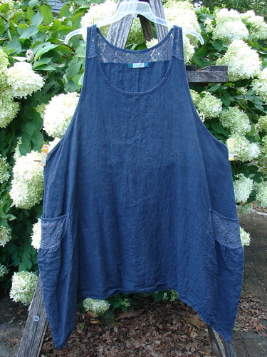 Barclay Summercloth Lace Two Pocket Pinafore in unpainted black, size 2, shown as a loose sleeveless dress with a rounded neckline, lace-trimmed shoulder straps, and two front patch pockets. The textured fabric hangs in a soft A-line shape against leafy white hydrangeas and weathered wood.