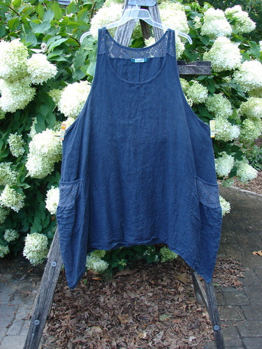 Barclay Summercloth Lace Two Pocket Pinafore in unpainted black, size 2, hangs from a wooden ladder outdoors. The loose, sleeveless dress has a rounded neckline, two side pockets with lace trim, and a soft, textured drape.