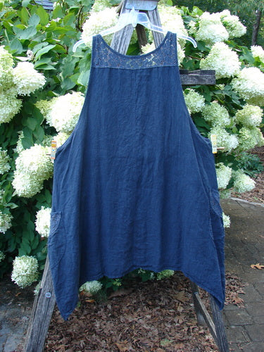 Barclay Summercloth Lace Two Pocket Pinafore in unpainted black, size 2, hangs from a wooden frame. The sleeveless dress has a lace-trimmed neckline, two side pockets, and a softly textured, draped fabric in deep blue-black.