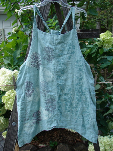 Barclay Linen Side Pocket Apron Jumper Fan Floral Dusty Sage Size 0 displayed on a wooden fence, featuring sectional panels, two side drop pockets, adjustable shoulder straps, and a continuous floral pattern.