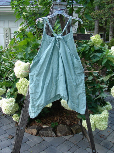 Barclay Linen Side Pocket Apron Jumper in Dusty Sage displayed on a wooden rack, featuring floral patterns and adjustable shoulder straps with sectional panels and wrap side drop pockets.