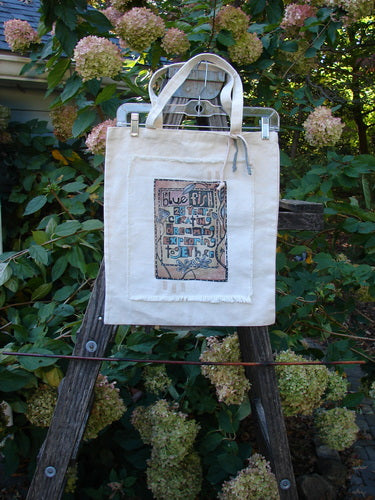 The 2000 PMU Patched Cotton Canvas Pocket Tote Promo Natural One Size is displayed on a wooden stand, featuring intricate patchwork and fringe accents, showcasing its unique upcycled design from Bluefishfinder.com.