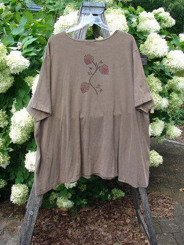 Barclay Short Sleeved Studio Tee Tree Offer Fauna Size 3 in muted taupe, a relaxed short-sleeve tee with a loose drape and rounded neckline. A small branch-and-berry motif sits at the center chest, while the soft cotton fabric hangs from a clear hanger against dense white hydrangeas and green foliage.