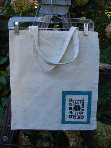 2000 PMU Patched Cotton Canvas Pocket Tote Promo Natural One Size hanging on a swinger, showcasing blue square designs, tote length shoulder straps, and a signature Blue Fish patch with fringe accents.