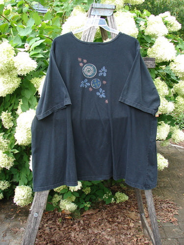 Barclay Short Sleeved Studio Tee Magic Moon Cloud Black Size 3 in washed black cotton, hanging from a wooden easel outdoors. The loose short-sleeved tee has a round neck and a small celestial graphic centered on the chest, with soft light and pale flowers behind it.