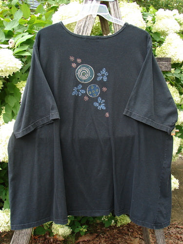 Barclay Short Sleeved Studio Tee Magic Moon Cloud Black Size 3, a black short-sleeved tee with a loose drape and a centered moon-and-cloud graphic in soft blue, teal, and pink. The cotton-like knit hangs from a hanger outdoors, with leafy greenery framing the dark fabric.
