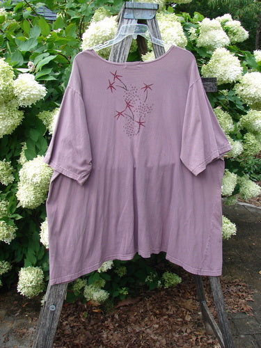 Barclay Short Sleeved Studio Tee Tree Star Laurel Size 3, a loose mauve short-sleeved T-shirt with a round neckline and a small tree star laurel graphic on the chest. The soft knit drapes in a boxy shape, hanging from a wooden stand against white hydrangeas and green foliage.