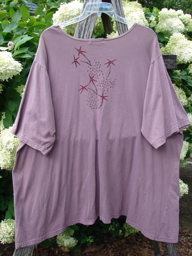 Barclay Short Sleeved Studio Tee Tree Star Laurel Size 3 in muted laurel purple, a loose short-sleeved T-shirt with a wide scoop neckline and draped hem. The front shows a delicate tree-star graphic in burgundy and gray, displayed on a hanger against leafy greenery and white blossoms.