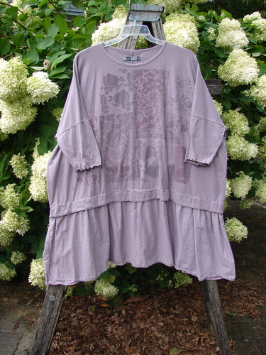 Barclay Batiste Banded Flounce Esperanza Tunic in Strawberry Garden Sea Fog, a loose sea-fog lavender top with a faded floral front print, banded neckline, elbow-length flounce sleeves, and a tiered, softly gathered hem. It hangs from a clear hanger on a weathered wooden ladder, framed by white hydrangeas and dense green foliage.