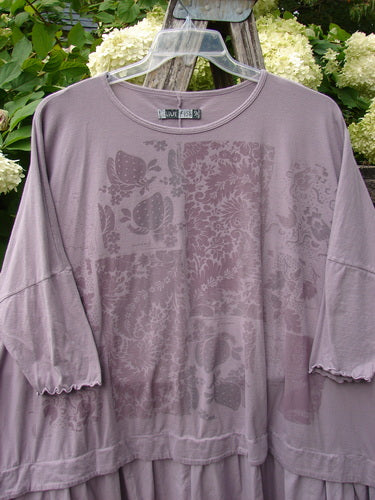 Barclay Batiste Banded Flounce Esperanza Tunic Strawberry Garden Sea Fog OSFA in muted sea fog, a relaxed long-sleeve tunic with a round neckline, banded hem, and soft flounce trim. The lightweight fabric shows a faded floral-and-butterfly print, hanging on a clear hanger against greenery.
