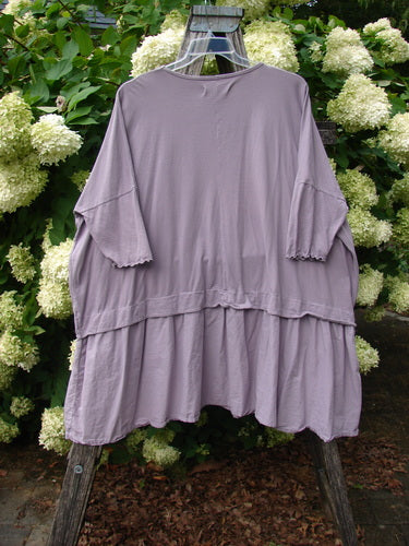 Barclay Batiste Banded Flounce Esperanza Tunic in Strawberry Garden Sea Fog, a loose lavender-gray pullover with wide three-quarter sleeves, gathered banded seams, and a layered flounce hem. The soft, draped fabric hangs from a rustic wooden ladder against white hydrangeas and green foliage.