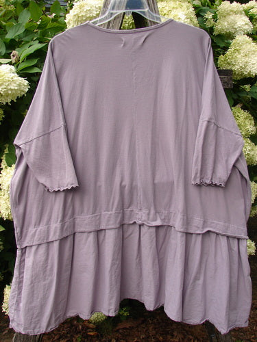 Barclay Batiste Banded Flounce Esperanza Tunic in Strawberry Garden Sea Fog, a loose lavender-gray batiste tunic with wide sleeves, a banded yoke, and a softly flounced hem. The lightweight fabric hangs in relaxed folds, with the garment centered against leafy white hydrangeas.