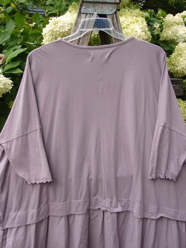 Barclay Batiste Banded Flounce Esperanza Tunic in Sea Fog, a loose, mauve-gray top with a wide boat neckline, elbow-length flounce sleeves, and a tiered ruffled hem. Light fabric drapes softly against the outdoor greenery backdrop.