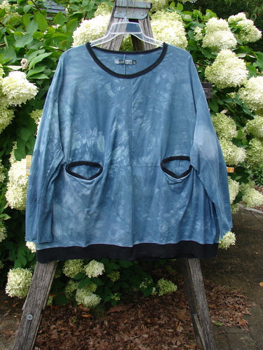 Barclay Cotton Lycra Open Rib Pocket Pullover in unpainted mottled blue shore, OSFA, with a loose drape, open rib texture, black trim at the neckline and hem, and two oval front pockets. It hangs on a wooden stand amid white hydrangeas and green foliage.
