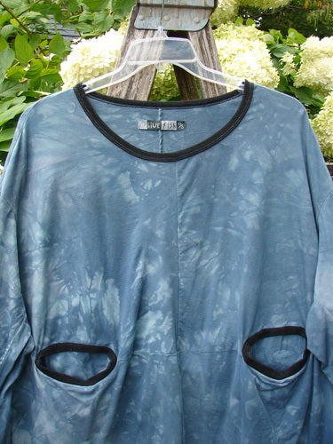 Barclay Cotton Lycra Open Rib Pocket Pullover in Unpainted Mottled Blue Shore OSFA, a loose long-sleeve top with a black ribbed neckline and oval pocket openings trimmed in black. The mottled blue fabric hangs on a clear hanger against leafy greenery and pale flowers.
