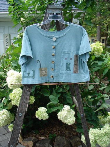 1994 Song Top Multi Letter K Ice Size 1: A blue shirt with a wide boxy shape, shallow neckline, vintage buttons, and a painted front pocket, displayed on a swinger.