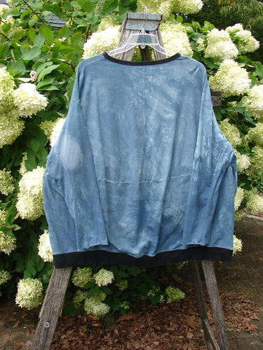 Barclay Cotton Lycra Open Rib Pocket Pullover in unpainted mottled blue shore, an OSFA top with a loose, draped silhouette, open rib texture, and dark trim at the neckline and hem, hanging on a wooden ladder outdoors among white hydrangeas.
