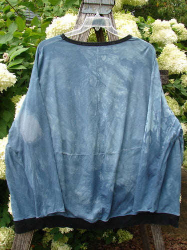 Barclay Cotton Lycra Open Rib Pocket Pullover in unpainted mottled blue shore, an oversized long-sleeve top with a sheer ribbed cotton-lycra body, black trim at the neckline and hem, and a softly draped, boxy silhouette hanging on a clear hanger amid pale hydrangeas.