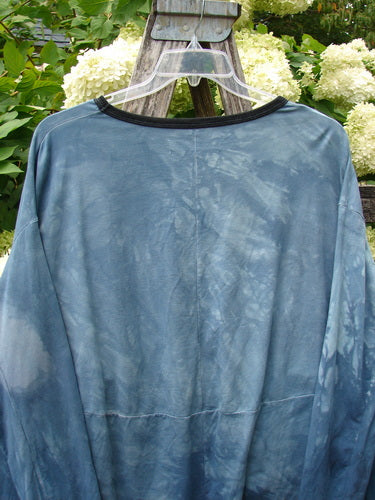 Barclay Cotton Lycra Open Rib Pocket Pullover in unpainted mottled blue shore, an OSFA top with a loose drape, dark ribbed crew neckline, and soft cotton-lycra texture. It hangs on a clear hanger against leafy hydrangeas and weathered wood.