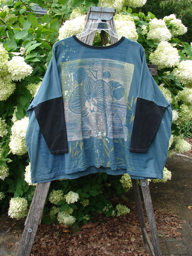 Barclay Cotton Lycra Rib Sleeved Treesoul Pullover Orchid Forest OSFA in blue-green cotton Lycra, with black ribbed sleeves and a loose, boxy silhouette. The front shows a large floral graphic, and it hangs on a wooden ladder amid white blossoms and green foliage.