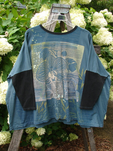 Barclay Cotton Lycra Rib Sleeved Treesoul Pullover Orchid Forest OSFA in blue-green with black ribbed sleeves and collar, featuring a large faded floral graphic across the front. The loose knit top hangs from a wooden ladder amid white hydrangeas, with soft outdoor light deepening the muted forest tones.