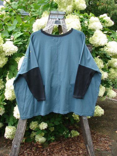 Barclay Cotton Lycra Rib Sleeved Treesoul Pullover in Orchid Forest, an OSFA teal-blue top with black ribbed sleeves and a black crew neckline. It hangs from a wooden ladder against dense white hydrangea blooms, showing a relaxed, boxy drape and soft cotton-lycra texture.