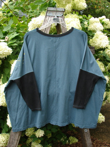 Barclay Cotton Lycra Rib Sleeved Treesoul Pullover Orchid Forest OSFA in muted blue-green cotton Lycra, with black ribbed long sleeves and a black crew neckline. The loose pullover hangs on a wooden ladder amid pale hydrangeas and leafy greenery.