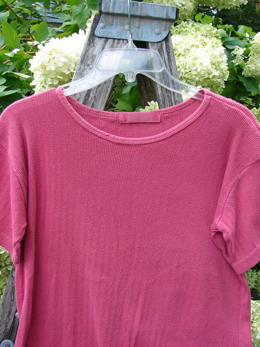 1997 Pointelle Short Sleeved Undertop in unpainted geranium, Tiny Size 2, a ribbed pink short-sleeve top with a round neckline and fine pointelle texture. It hangs on a clear hanger against weathered wood and soft white hydrangeas, with natural greenery behind.