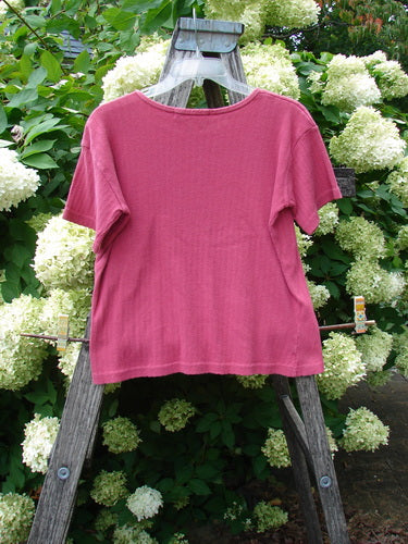 1997 Pointelle Short Sleeved Undertop in unpainted geranium, tiny size 2, a pink short-sleeve top with a fine pointelle texture and simple crew neckline. It hangs centered on a weathered wooden ladder amid dense white hydrangea blooms and green foliage.