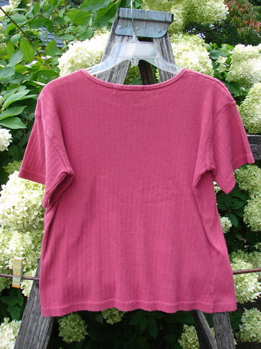 1997 Pointelle Short Sleeved Undertop in unpainted geranium, tiny size 2, a pink short-sleeved knit with a fine pointelle texture, gently scooped neckline, and softly flared hem, draped on a wooden hanger against pale hydrangeas and green foliage.