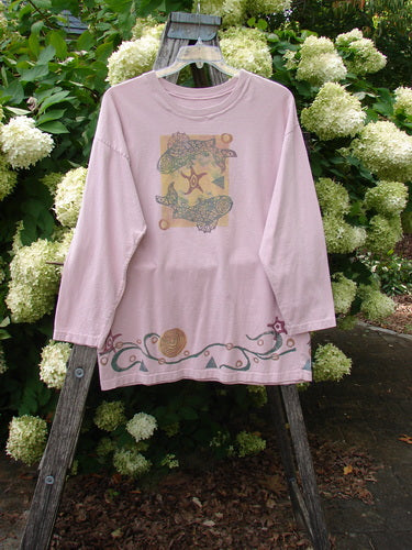 1993 Long Sleeved Tee Ocean Folly Ask Pink OSFA in soft pink cotton, hanging from a wooden ladder outdoors. The long sleeves and crew neck frame a faded graphic print on the chest and a decorative border near the hem, with white hydrangeas and green foliage behind it.