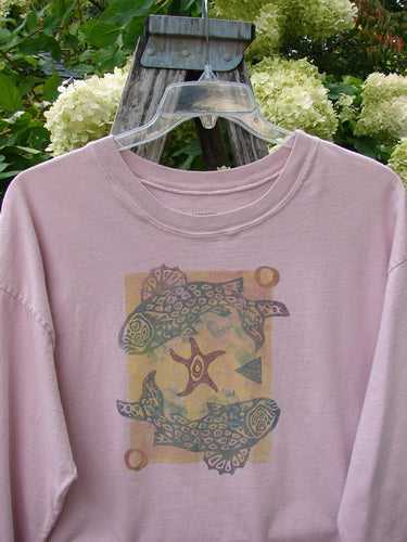 1993 Long Sleeved Tee Ocean Folly Ask Pink OSFA in soft pink cotton, with a relaxed crew neck and long sleeves. The front shows a faded ocean-inspired graphic of two fish-like forms around a central star motif, hanging on a clear hanger against green foliage and pale blossoms.